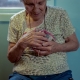 An elderly woman wearing a beige coloured t-shirt cradles unidentified objects against her chest with both hands. She is smiling and looks down toward the objects.  