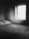 Black and white photograph of a bed underneath a window with a ghostly presence hovering above it.