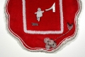 Red felt in an organic shape embroidered with white beads and flower imagery. In the middle is a black-and-white photograph of a baby reaching for an adult’s hand. 