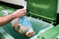 A hand carrying a bag filled with food waste about to be dropped into a green bin. / Une main dépose un sac rempli de déchets alimentaires dans un bac vert. 
