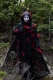une figure qui est vêtue d’une robe noire avec des détails rouges et a la peinture blanche sur leur visage. Une forêt en arrière-plan.