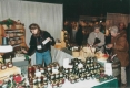 An artisan selling jams and other jarred items to  customers from behind a well-stocked table.