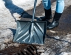 A close-up of a person wearing black rubber boots clearing snow away from a catch basin using a black shovel.