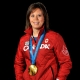 Headshot of Coach Carla MacLeod with an Olympic gold medal. She is wearing a red zip-up sweater with “Canada” written across the chest. 