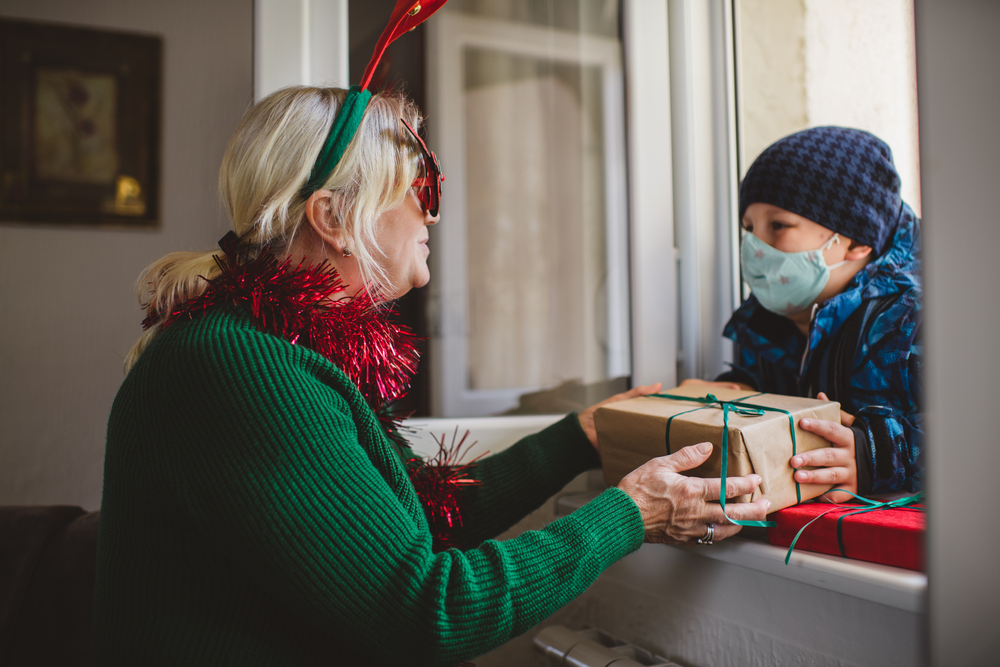An older white woman wearing antlers on her head and a red garland around her neck stands at an open window where her young grandson, wearing a mask, is passing her a wrapped package. 