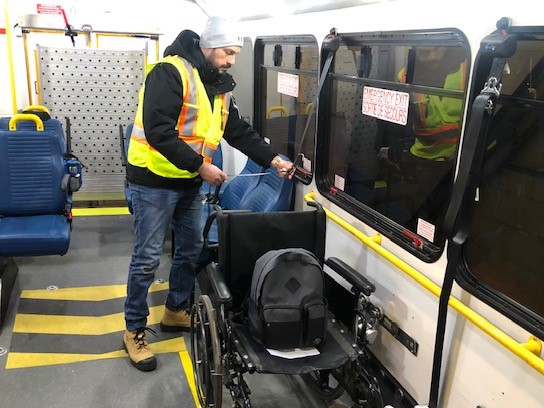 Un homme en formation vêtu d’un bonnet gris et d’un gilet de sécurité s’exerce à attacher un fauteuil roulant dans un autobus de Para Transpo. 