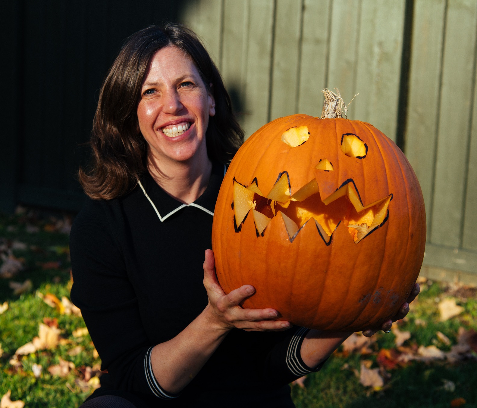 A woman with brown hair wearing a black shirt holds a large carved pumpkin in a yard with leaves on the ground on a sunny day. 