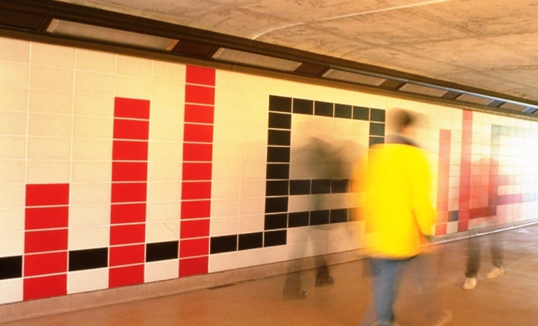 Photo de la murale le long du quai du Transitway composée de tuiles de béton rouges, blanches et noires disposées dans un motif géométrique. Des passants révèlent l’ampleur de l’œuvre.