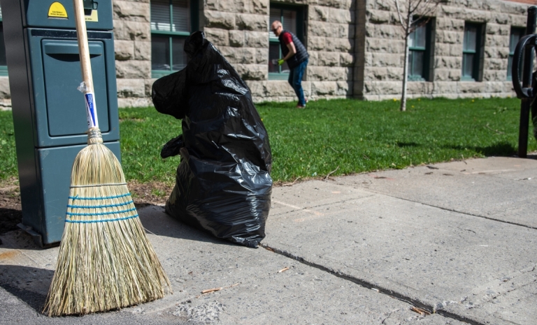 A broom and a half full plastic garbage bag on the sidewalk with a man picking up litter in the background