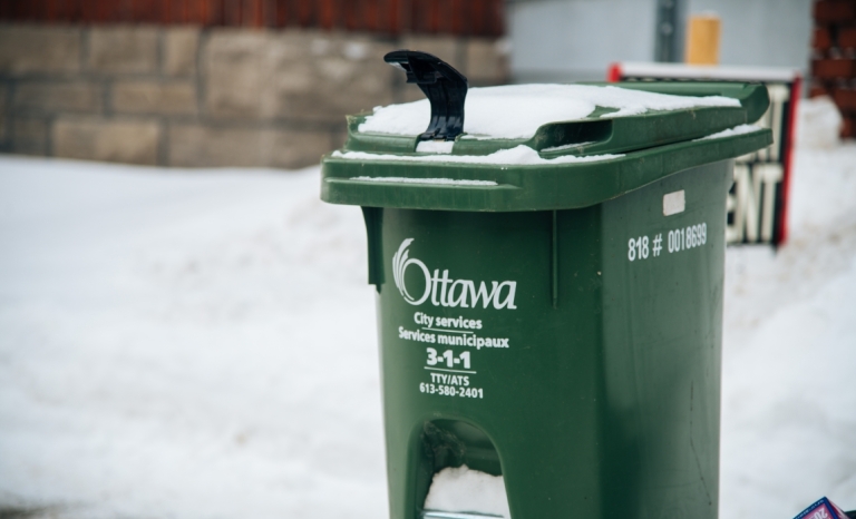 A compost bin on the sidewalk with an "Ottawa" logo in the winter