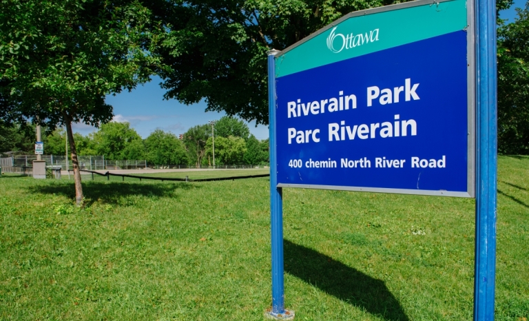 A sign for Riverain Park with a baseball diamond in the background