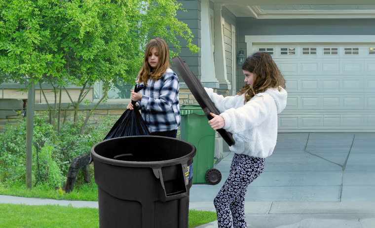 Deux enfants en train de déposer un sac d’ordures dans un bac à déchets./ Two kids tossing a garbage bag into a garbage bin.