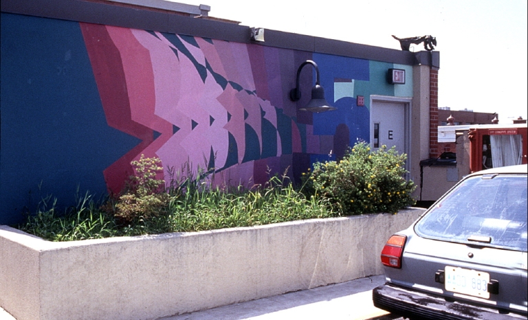 Mural of geometric shapes in pink, purple and red with blue background on the side of a building near a parking lot