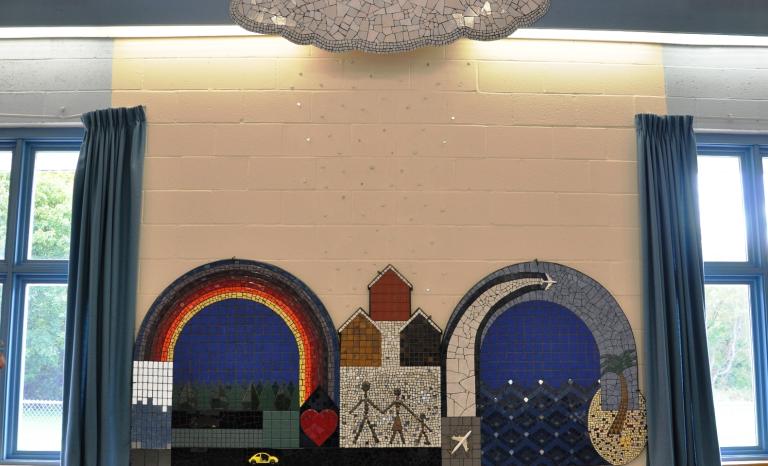 Image of the primarily mosaic tile sculpture showing components of the community: forests, water, planes, cars, love a rainbow, a beach and a family. 