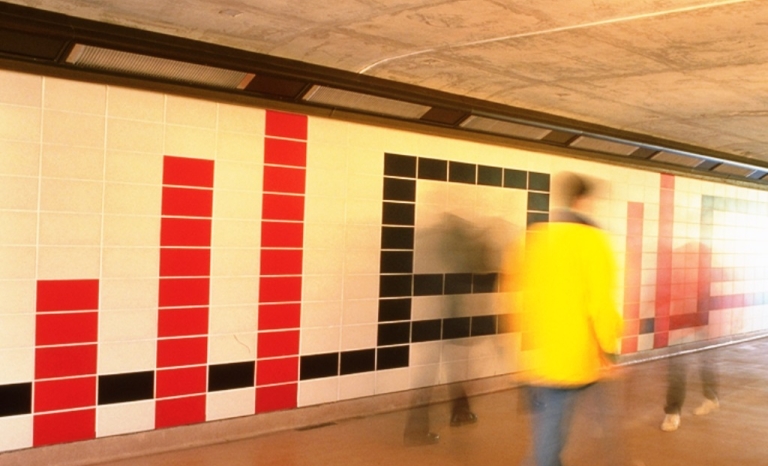 An image of the mural at the transitway platform composed of red, white and black concrete tiles arranged in a geometric pattern. Passersby help reveal the scale of the work.