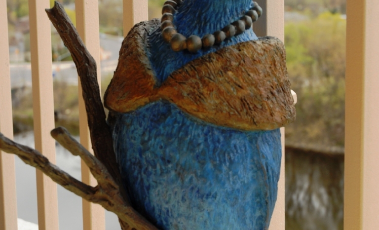 A sculpture of a blue bird chirping while sitting on two branches wearing a pearl necklace and a fur wrap and hat.