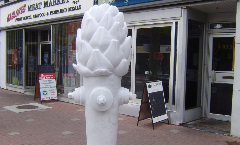 Image shows a sculpture of an oversized artichoke on top of a fire hydrant base and mounted on a plinth. 