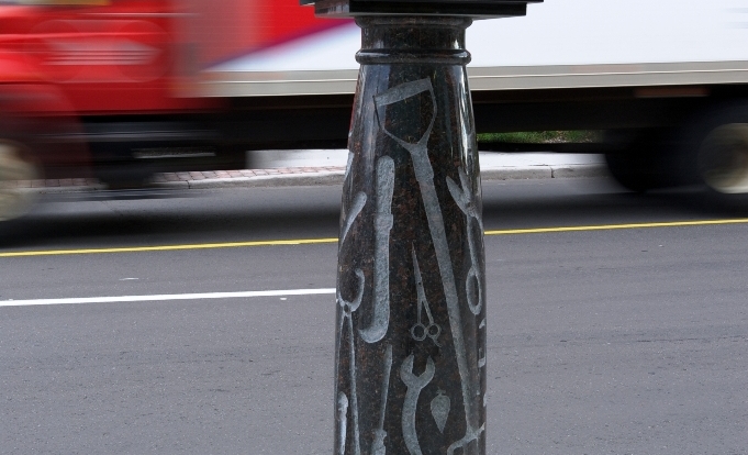 A bronze hat, parcel and envelope rest on a tray atop a marble column carved with silhouettes of the tools used in trades.