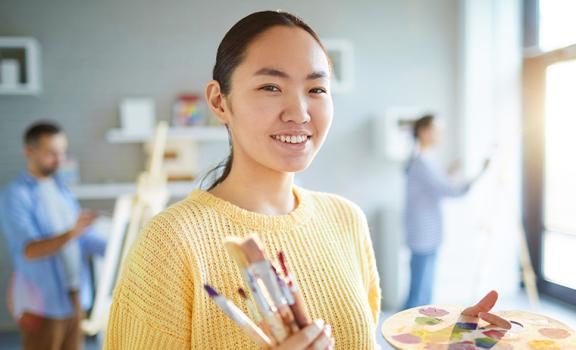 Camera focuses on a woman in an art studio painting. Other painters are in the background.
