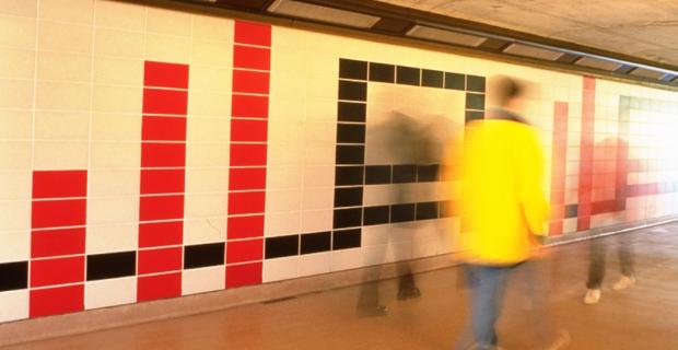 Photo de la murale le long du quai du Transitway composée de tuiles de béton rouges, blanches et noires disposées dans un motif géométrique. Des passants révèlent l’ampleur de l’œuvre.