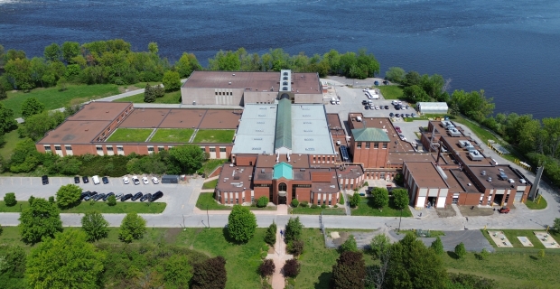Aerial view of a large brown brick facility surround by trees, located on the water.
