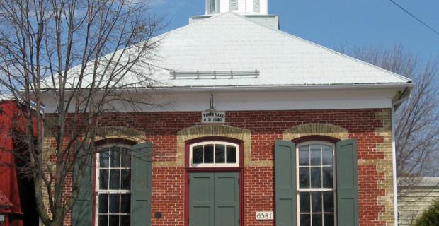 The exterior of a small red brick building hall with green doors and shudders and a white roof. 