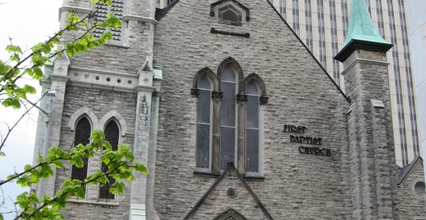 Church with limestone exterior and large wood front doors sits surrounded by tall office buildings downtown Ottawa.