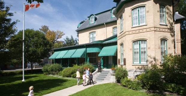 Un groupe de visiteurs se dirige vers un bâtiment de deux étages en briques beiges avec un toit gris et des détails verts.