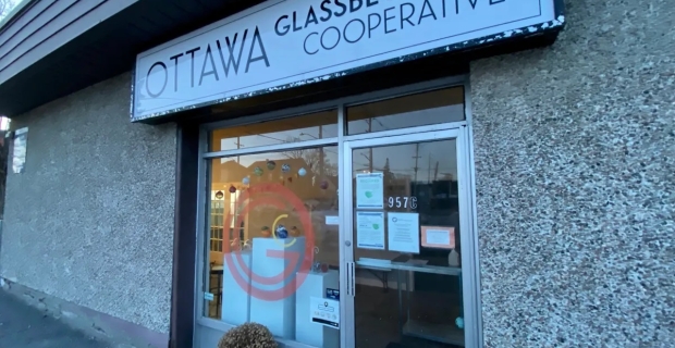 Un bâtiment gris avec une grande enseigne pour Ottawa Glassblowing Cooperative. Une plante à fleurs dans un pot blanc est assise à côté d’une porte vitrée. 