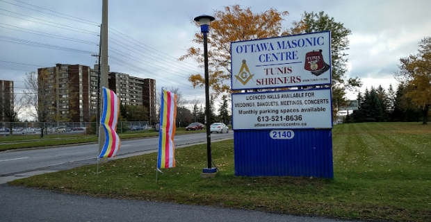 Une pancarte blanche pour le Centre maçonnique d’Ottawa sur un gradin bleu à côté d’un lampadaire. Les feuilles d’automne couvrent l’herbe derrière. 