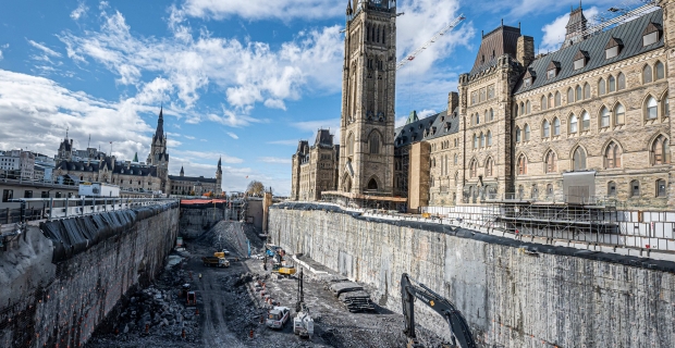 Large construction equipment in a deep excavation site beside Parliament building on a sunny day. 