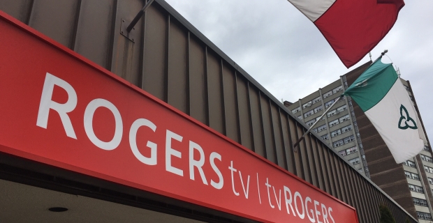 A red Rogers TV sign sits against a building with brown siding on a cloudy day. 