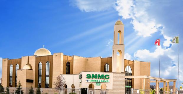 Exterior of a large beige and brown centre on a sunny day. A green and white sign in front reads" SNMC Centre & Masjid"