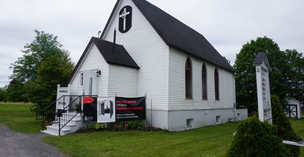 A white farmhouse church with black roof and three windows. 