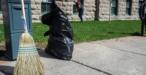 A broom and a half full plastic garbage bag on the sidewalk with a man picking up litter in the background