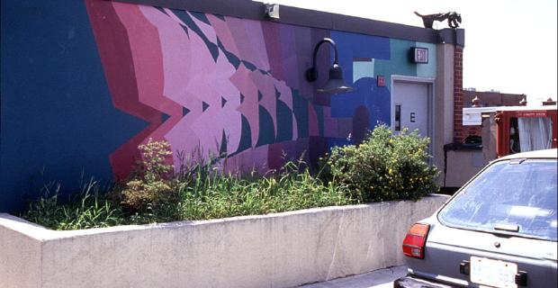 Mural of geometric shapes in pink, purple and red with blue background on the side of a building near a parking lot