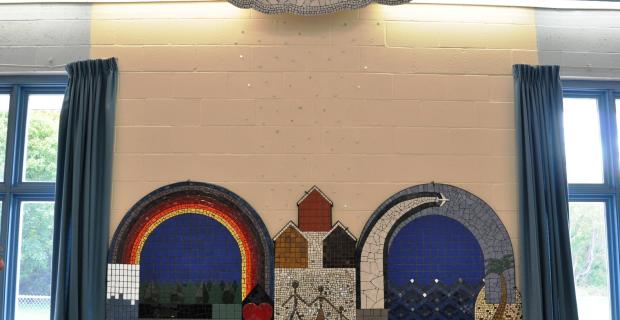 Image of the primarily mosaic tile sculpture showing components of the community: forests, water, planes, cars, love a rainbow, a beach and a family. 