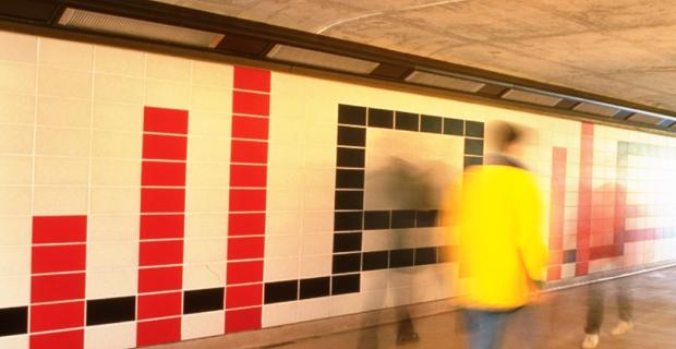 An image of the mural at the transitway platform composed of red, white and black concrete tiles arranged in a geometric pattern. Passersby help reveal the scale of the work.