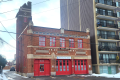 La façade extérieure en brique rouge, avec ses deux grandes portes à véhicules rouge pompier, sa porte piétonne et sa toiture surmontée d’une tour.