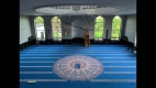 Salle de prière principale avec de grandes fenêtres, tapis bleu et lustre circulaire.