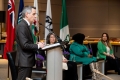 Le maire Sutcliff prononce un discours sur le podium, tandis que les conférenciers invités l’écoutent, assis.