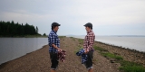 Two figures wearing plaid shirts, one blue, one red, face eachother. The setting is natural, with water on either side of the peninsula on which the figures stand.