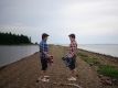 Two figures wearing plaid shirts, one blue, one red, face eachother. The setting is natural, with water on either side of the peninsula on which the figures stand.