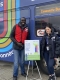 An OC Transpo male employee and a Safer Roads Ottawa female employee in front of an e-bus and a sign with information about large vehicle blind spots./Un employé d'OC Transpo et une employée de Sécurité des routes Ottawa devant un e-bus et un panneau d'information sur les angles morts des gros véhicules.