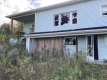 A blue two-storey house with broken windows and an open door, surrounded by long grass and weeds.