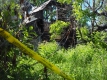 Ruines d’une maison brûlée autour desquelles la police a installé un ruban de sécurité. Le jardin est envahi par les broussailles et l’herbe haute.