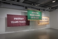 Three large textile artworks hang in a gallery space. They each have a text statement stitched on them.     