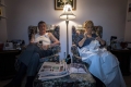 The image shows a couple sitting in their living room, the man with a remote and the woman sewing, enjoying a quiet evening together.