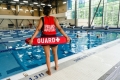 : A City of Ottawa lifeguard standing in front of a City swimming pool with people swimming in the background. 