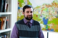 Portrait of a bearded Caucasian man wearing a purple shirt and a striped black and grey vest. Behind him are some books and a map of Ottawa with coloured blocks indicating zoning information. Portrait d’un homme blanc barbu portant une chemise violette et un gilet rayé noir et gris. Derrière lui, on aperçoit des livres et une carte d’Ottawa dotée de formes colorées représentant l’information sur le zonage. 